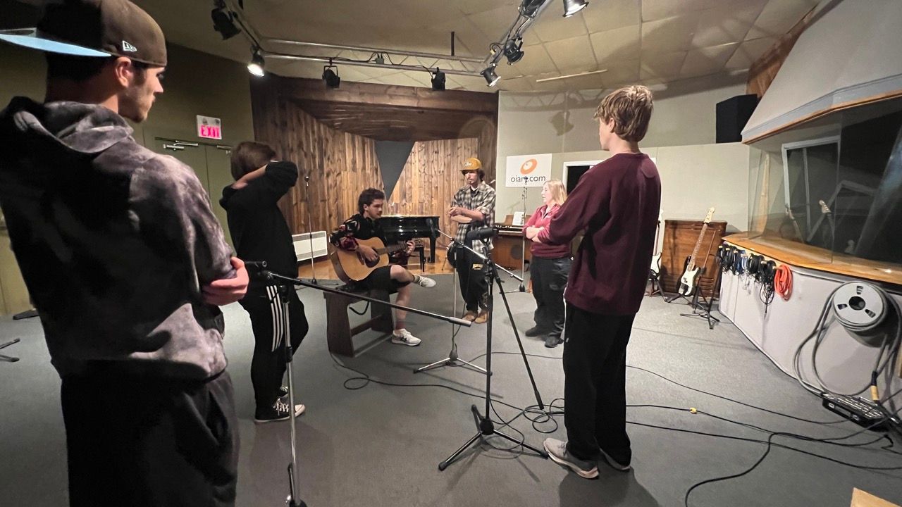 Band recording music in a studio. Guitarist sits, other members stand around, microphones present, and a window.