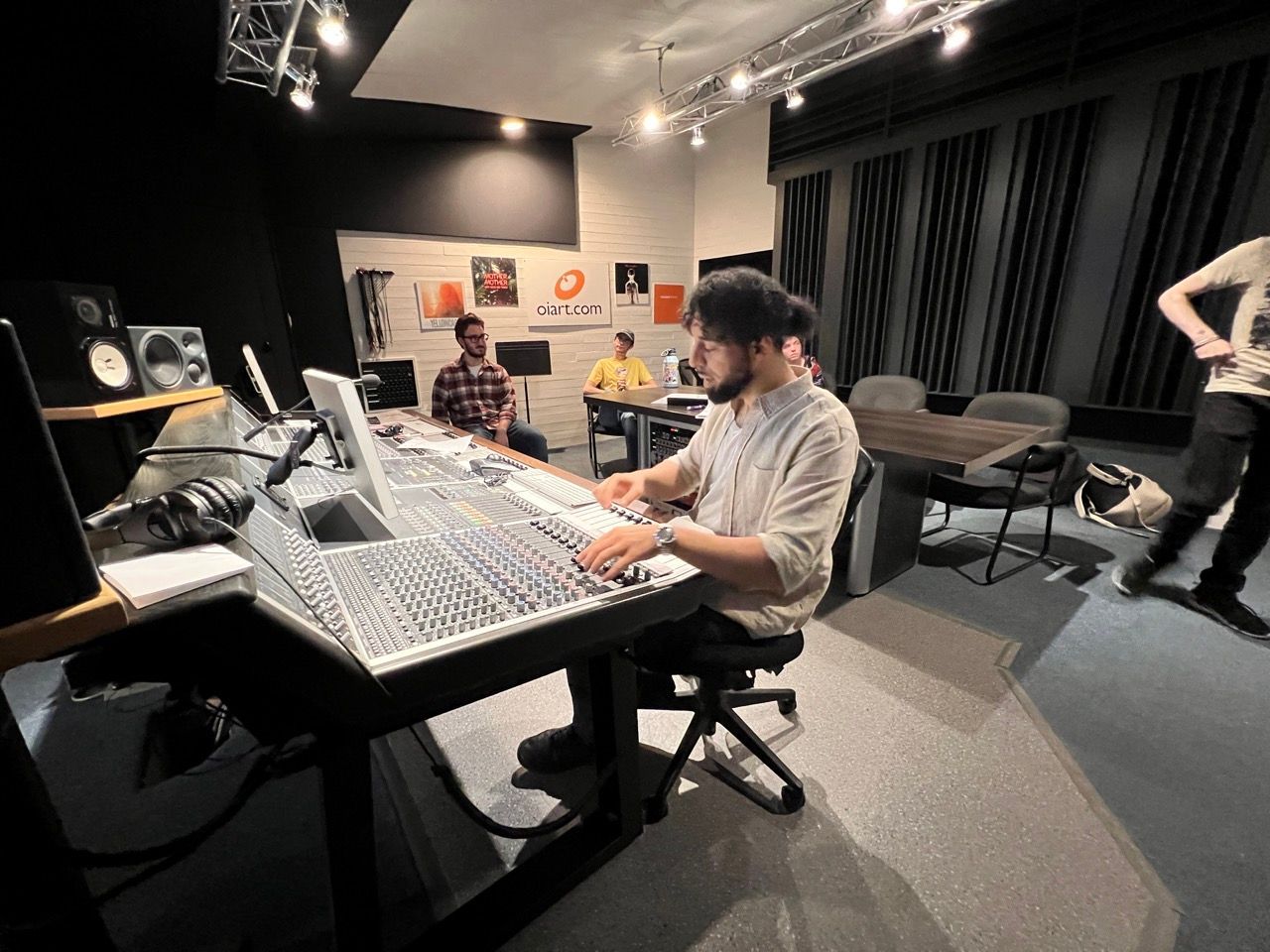Man operating audio equipment in a recording studio, with two other people seated nearby.