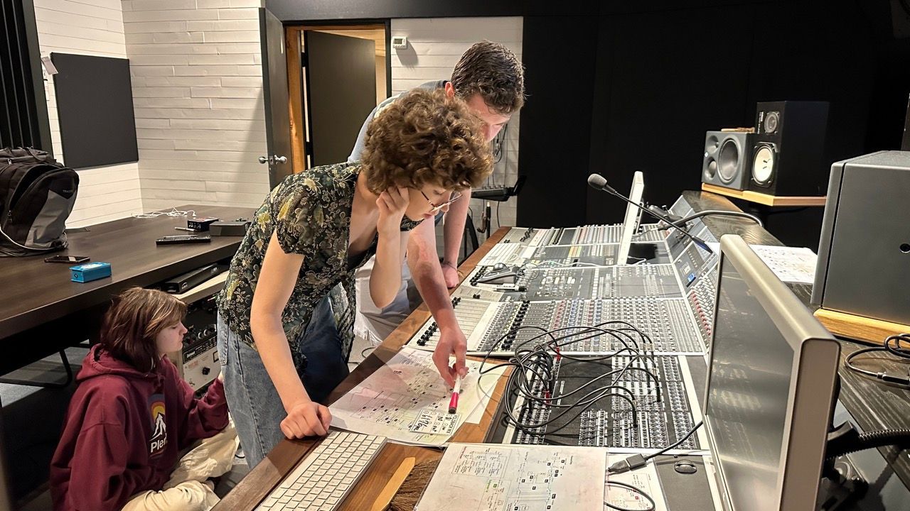 Three people in a recording studio examining a mixing board and papers. One points, while the others observe.