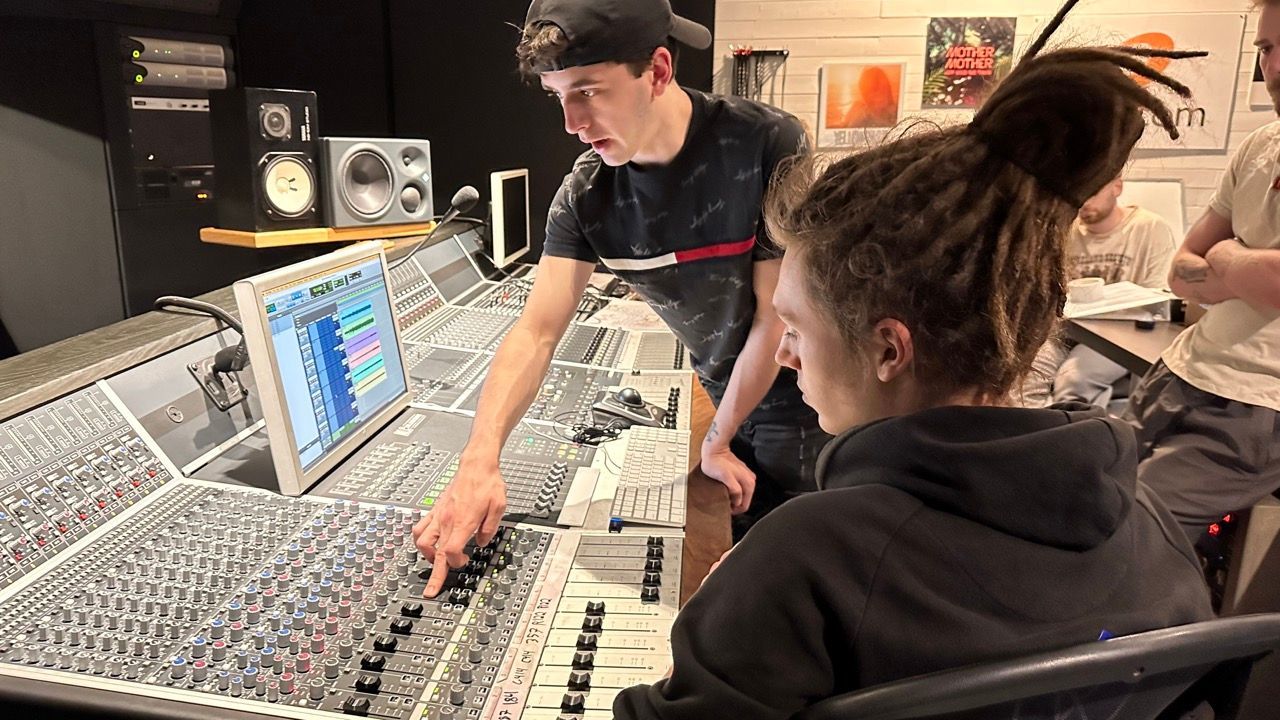 Two people in a recording studio adjust sound equipment, one pointing at a console.