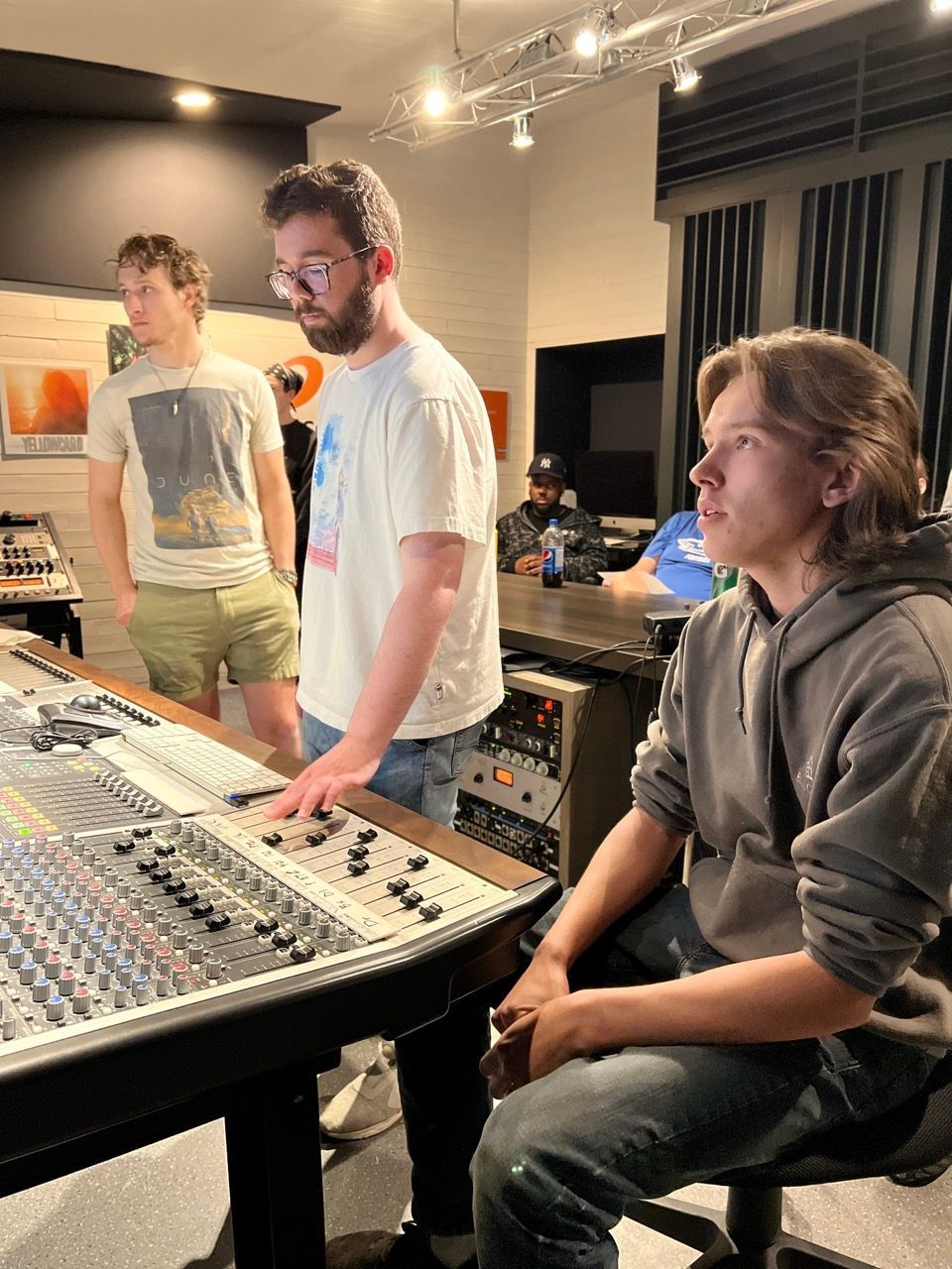 Three people in a recording studio; one at the soundboard, two observing. Dim lighting, mixing console.