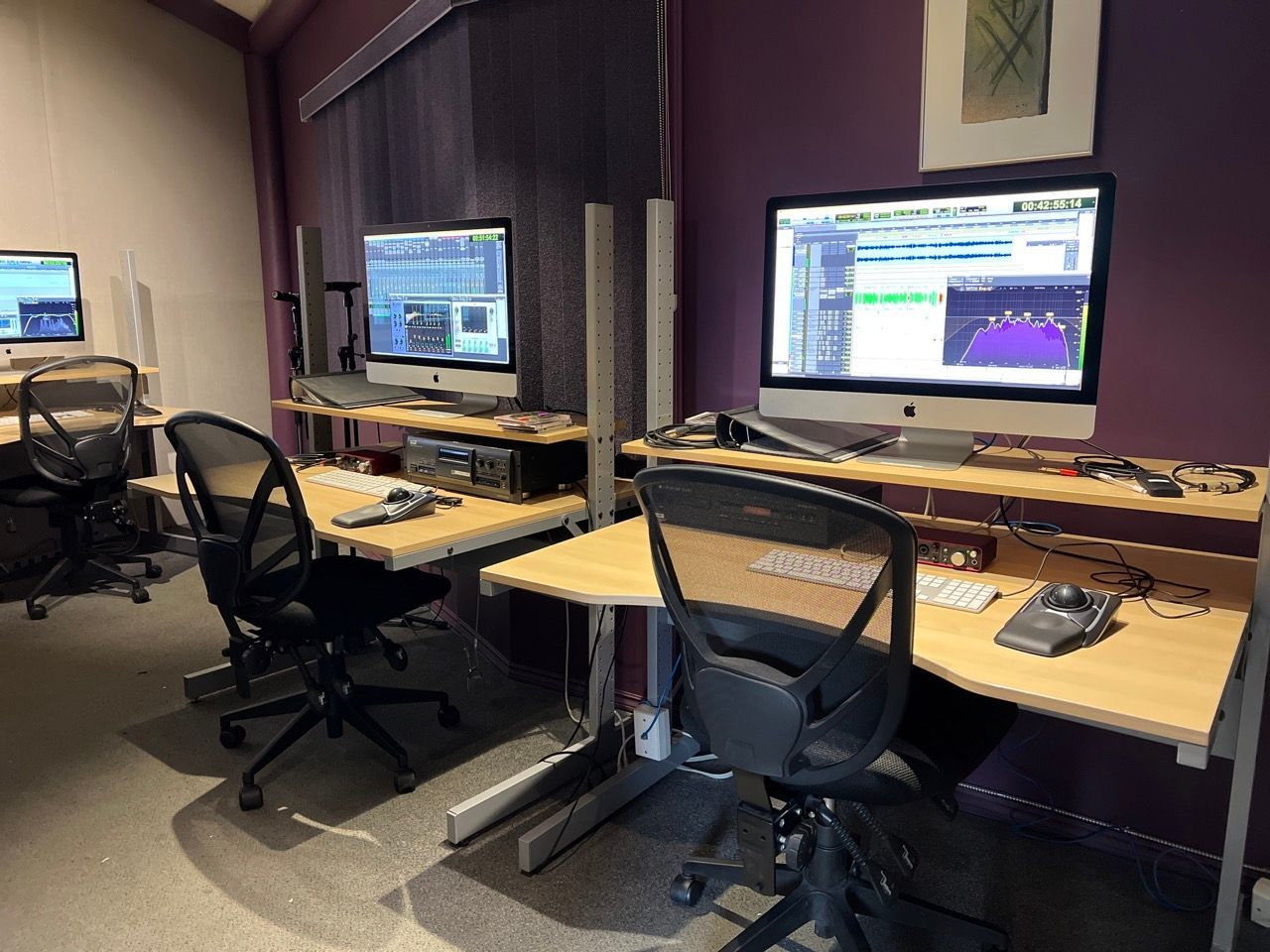 Recording studio with three computer workstations, monitors displaying audio editing software. Purple walls.