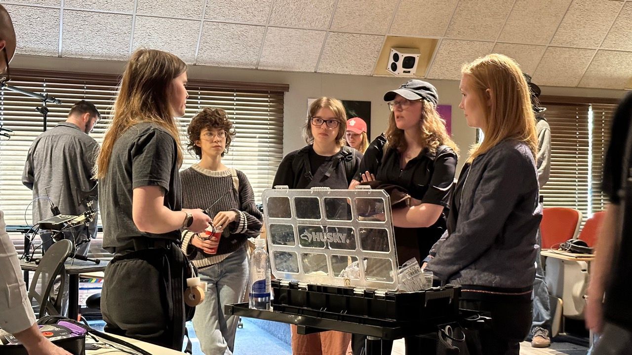 Group of students gathered around a table with a display. Woman explaining something, others listening intently. Indoors.