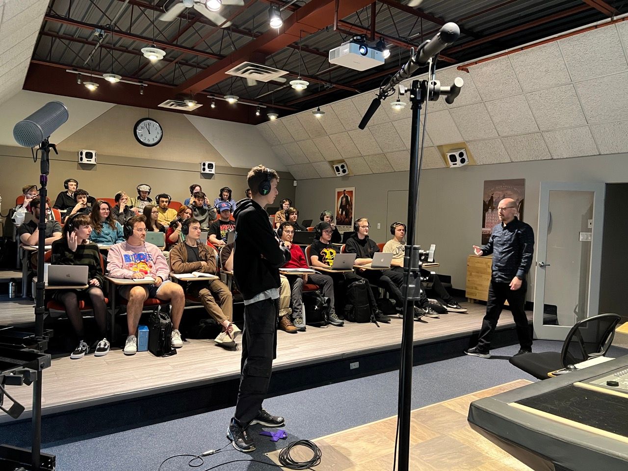 A man is standing in front of a microphone in front of a crowd of people in a classroom.