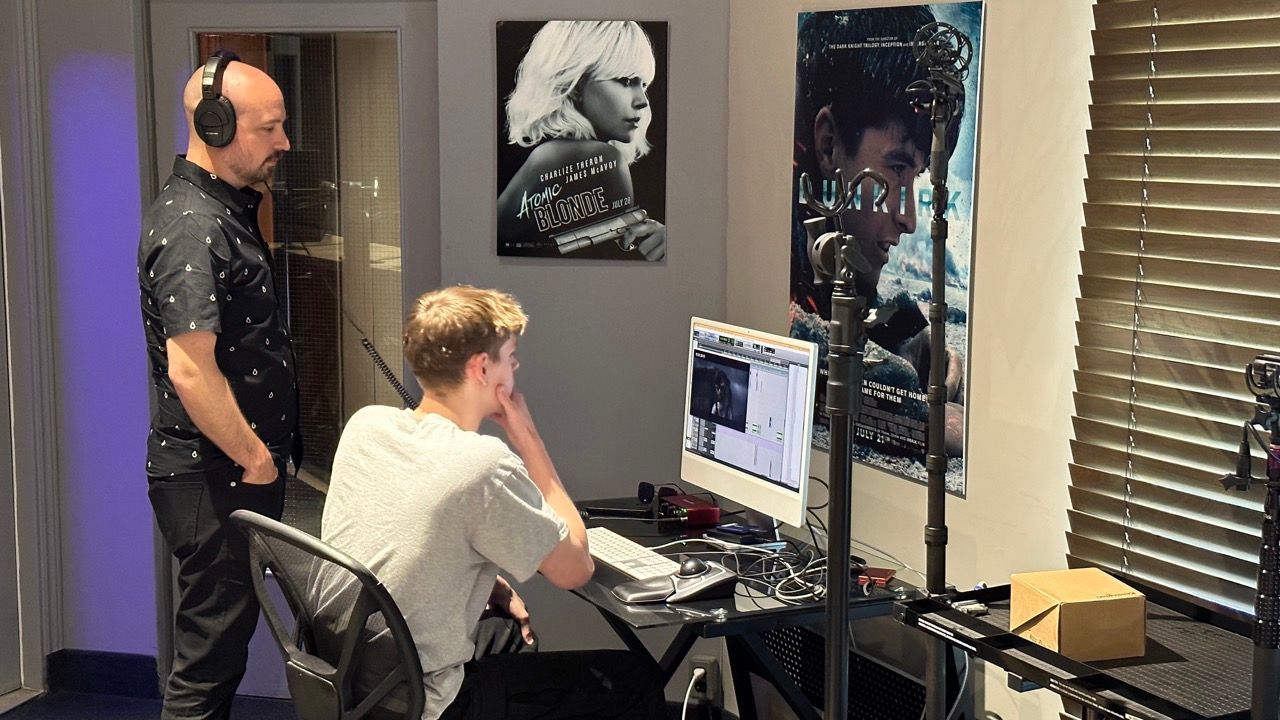 A man is standing next to a man sitting at a desk in front of a computer.