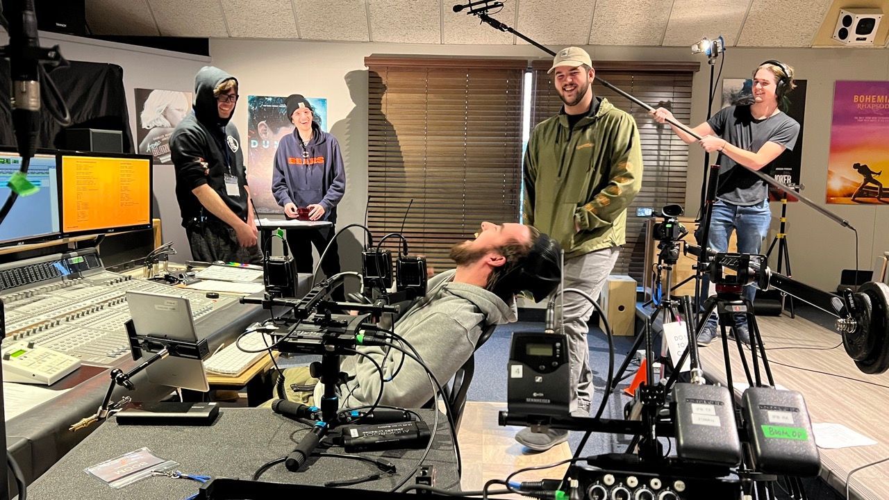 Film set: Crew members work around a man with his head tilted back. Equipment, monitors, and sound booms are present.