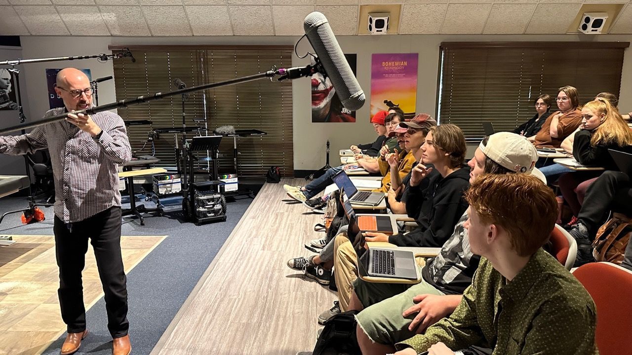 A man holds a microphone boom over a classroom of students, likely during a film production lecture.
