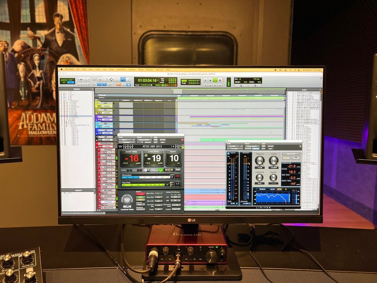 A computer monitor displays audio editing software, with an audio interface on a desk, and a poster behind it.