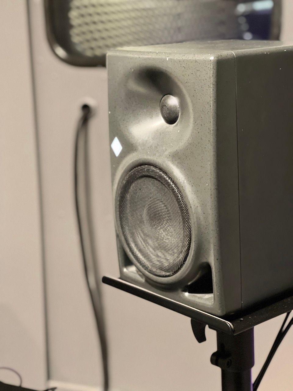 Gray studio monitor speaker on a stand with a rounded tweeter and woofer, in a recording studio.