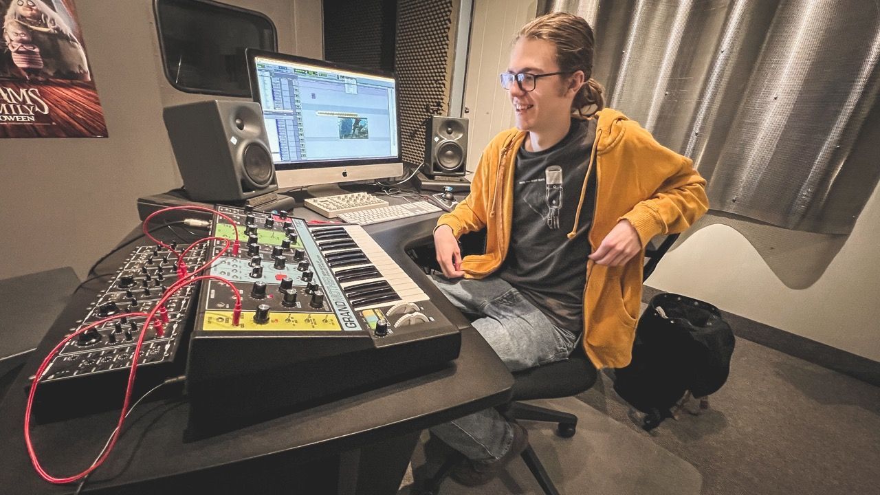 Person in a recording studio, seated at a desk with audio equipment. They wear glasses and a yellow hoodie.