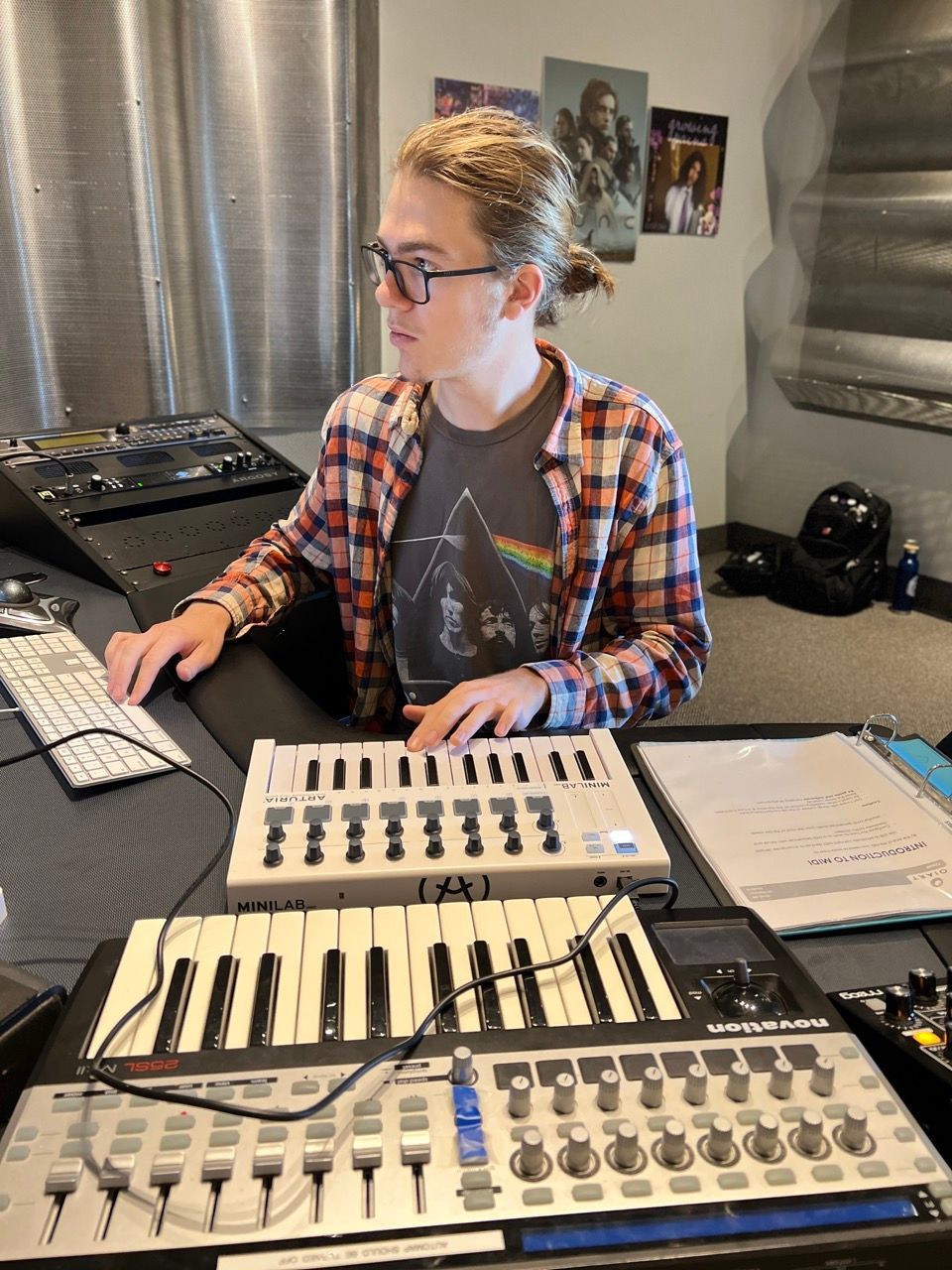 Person at a mixing console, using a keyboard and computer. They wear glasses and a plaid shirt, in a studio setting.