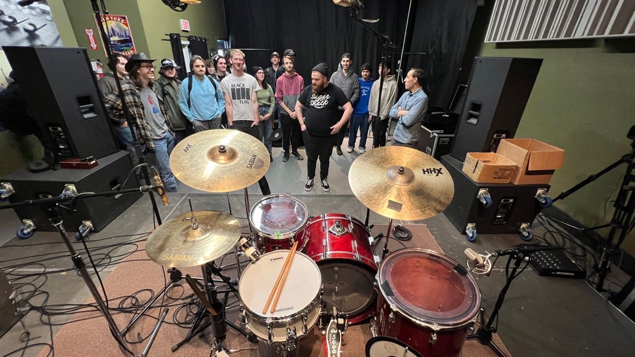 A group of people are standing around a drum set in a room.