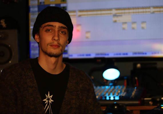Cam wearing a beanie and cardigan smiles in a music studio with equipment in the background.