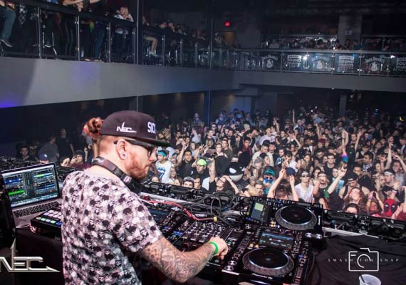 DJ at a crowded nightclub. Liam T with tattoos & hat at a console, people dancing with raised hands.