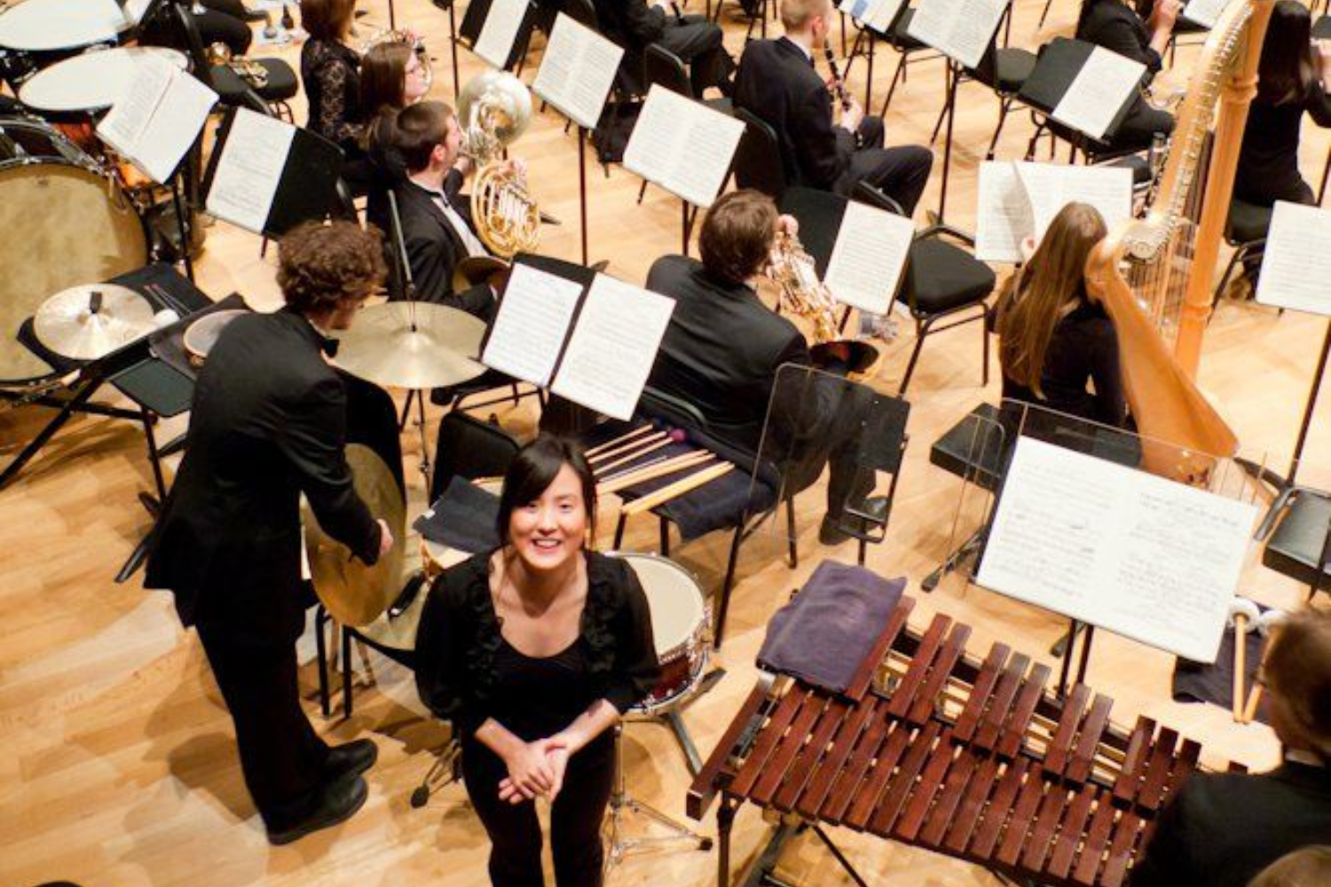 Orchestra rehearsal with musicians seated around a smiling woman in black near a marimba and harp