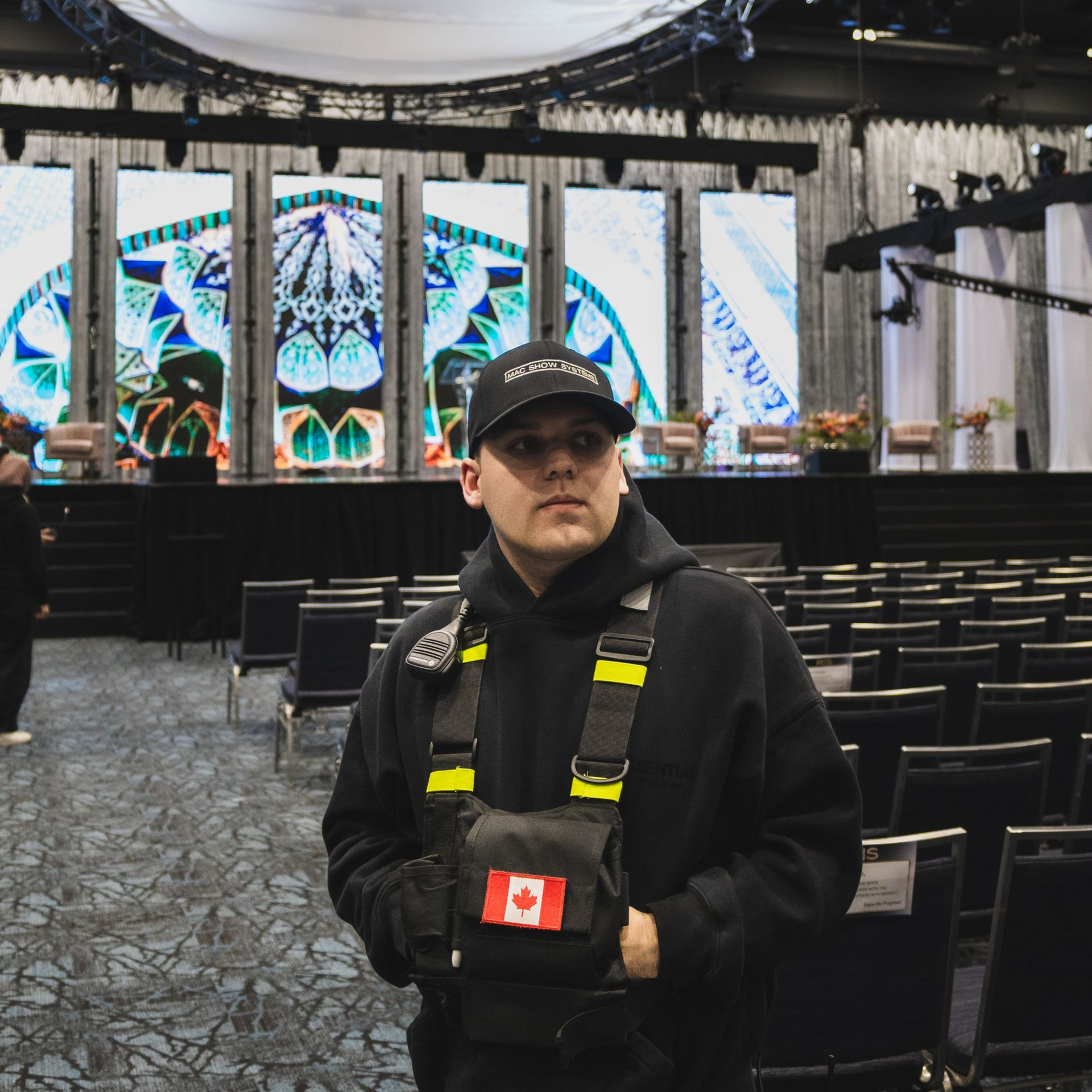 Avery Szumlanski in black jacket and Canadian flag gloves stands in an empty venue with illuminated stage screens.