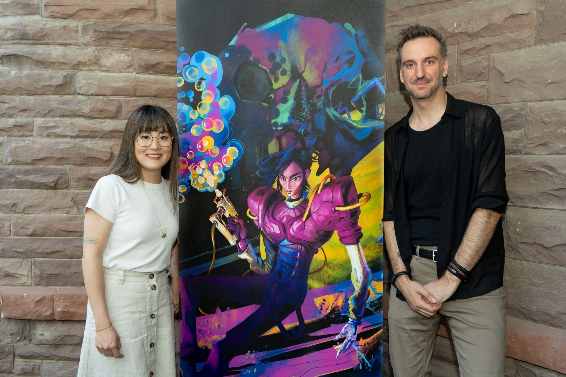 Two people standing beside a colorful neon superhero poster against a brick wall