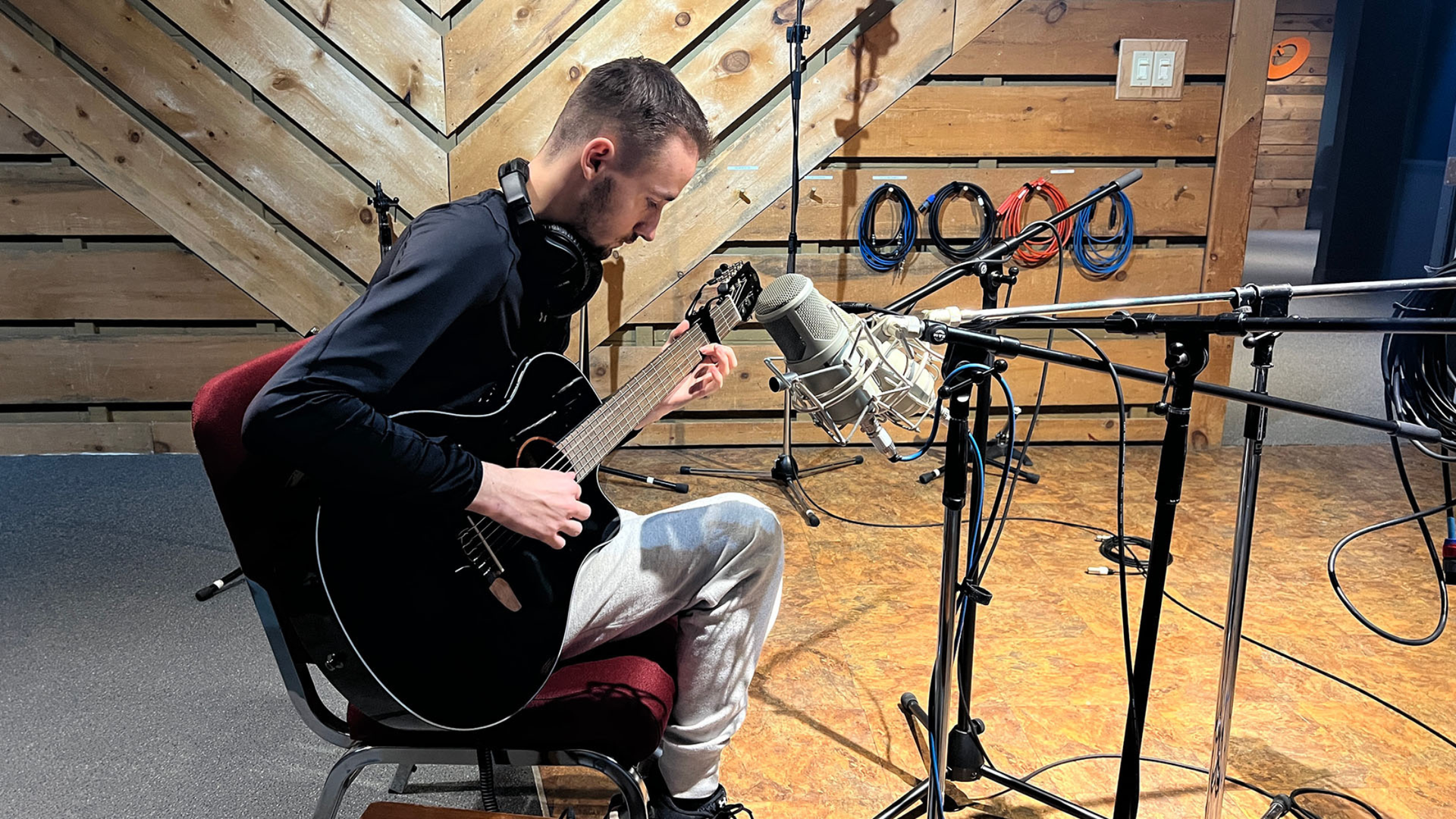 Man playing electric guitar in recording studio, seated. Microphone and headphones visible. Wooden wall in background.