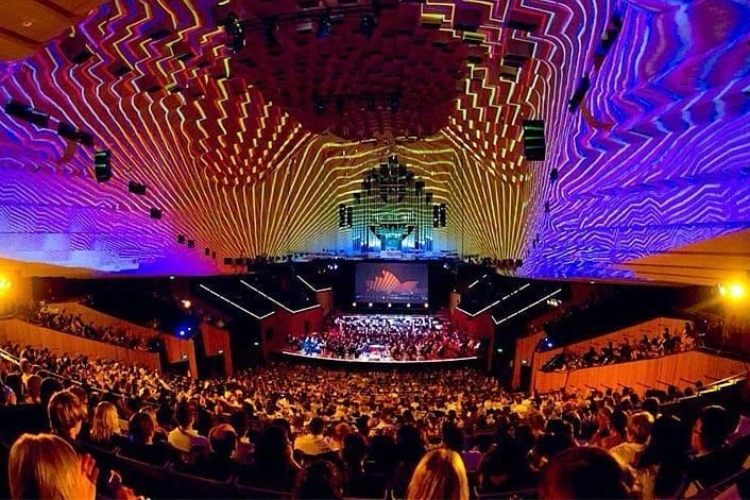 Crowded concert hall with a brightly lit stage and colorful, patterned ceiling lights