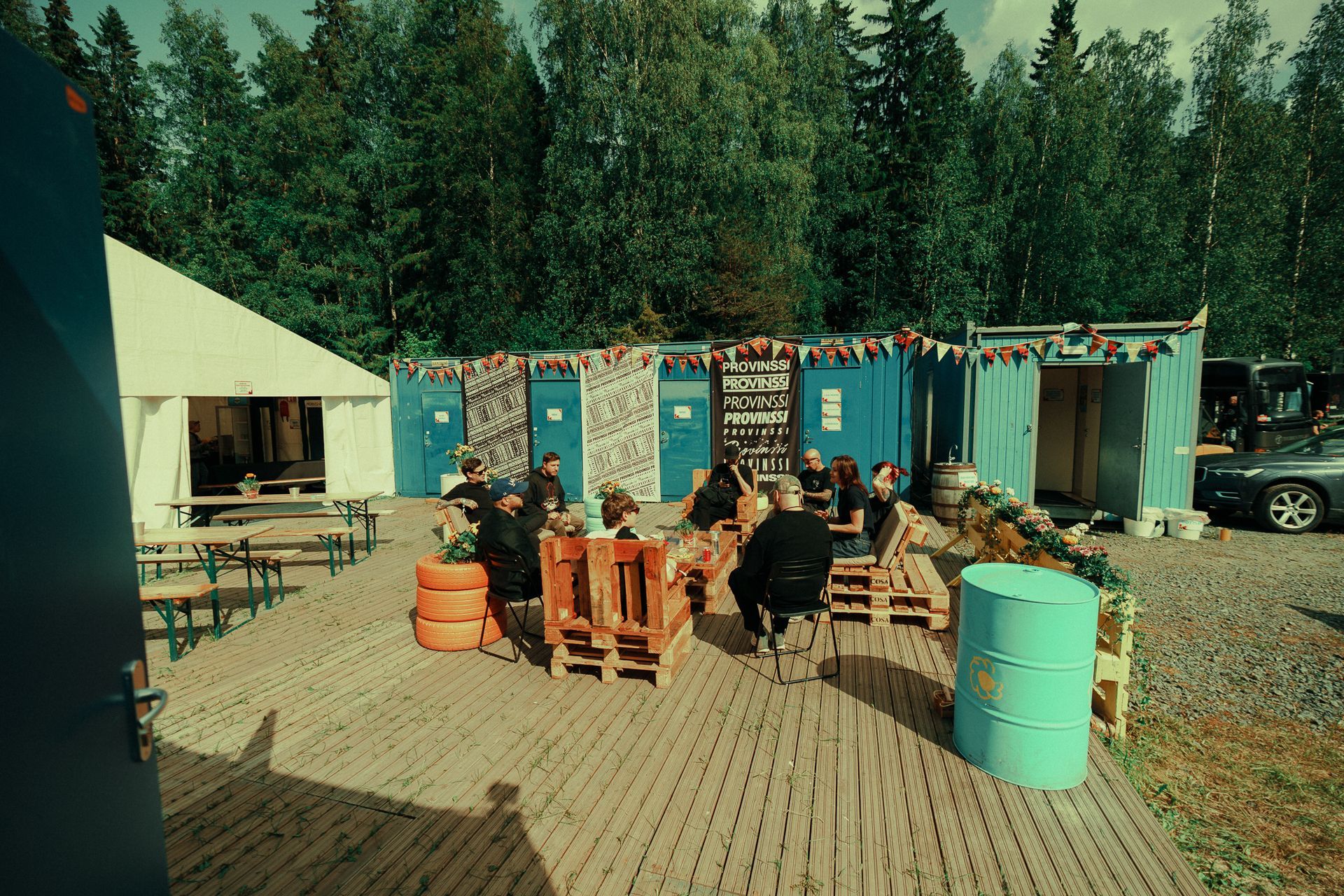 People seated outdoors at a rustic campsite with blue sheds and barrels in a wooded area.