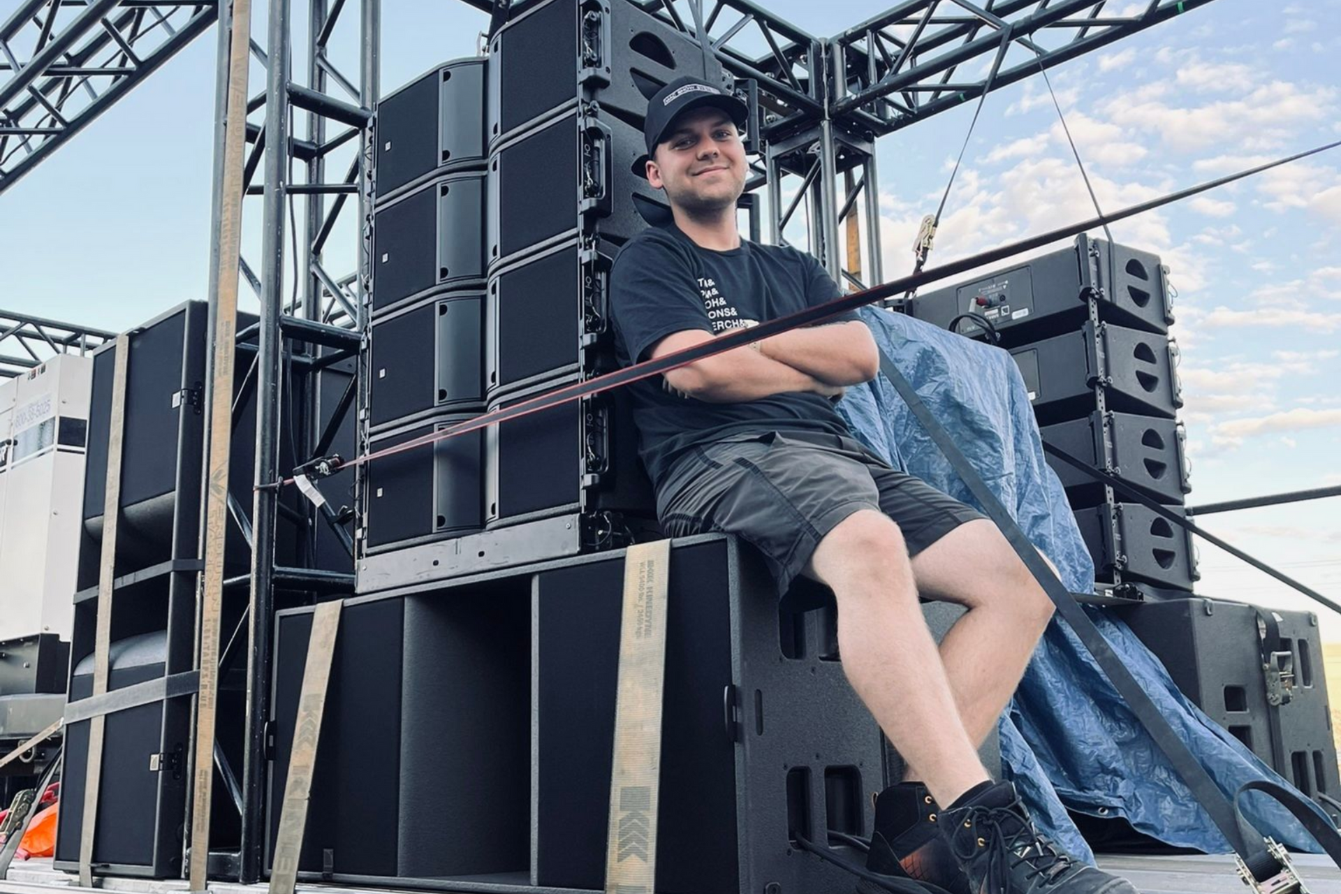 Avery Szumlamski sitting on stacked speakers at an outdoor stage, wearing a black shirt and cap.