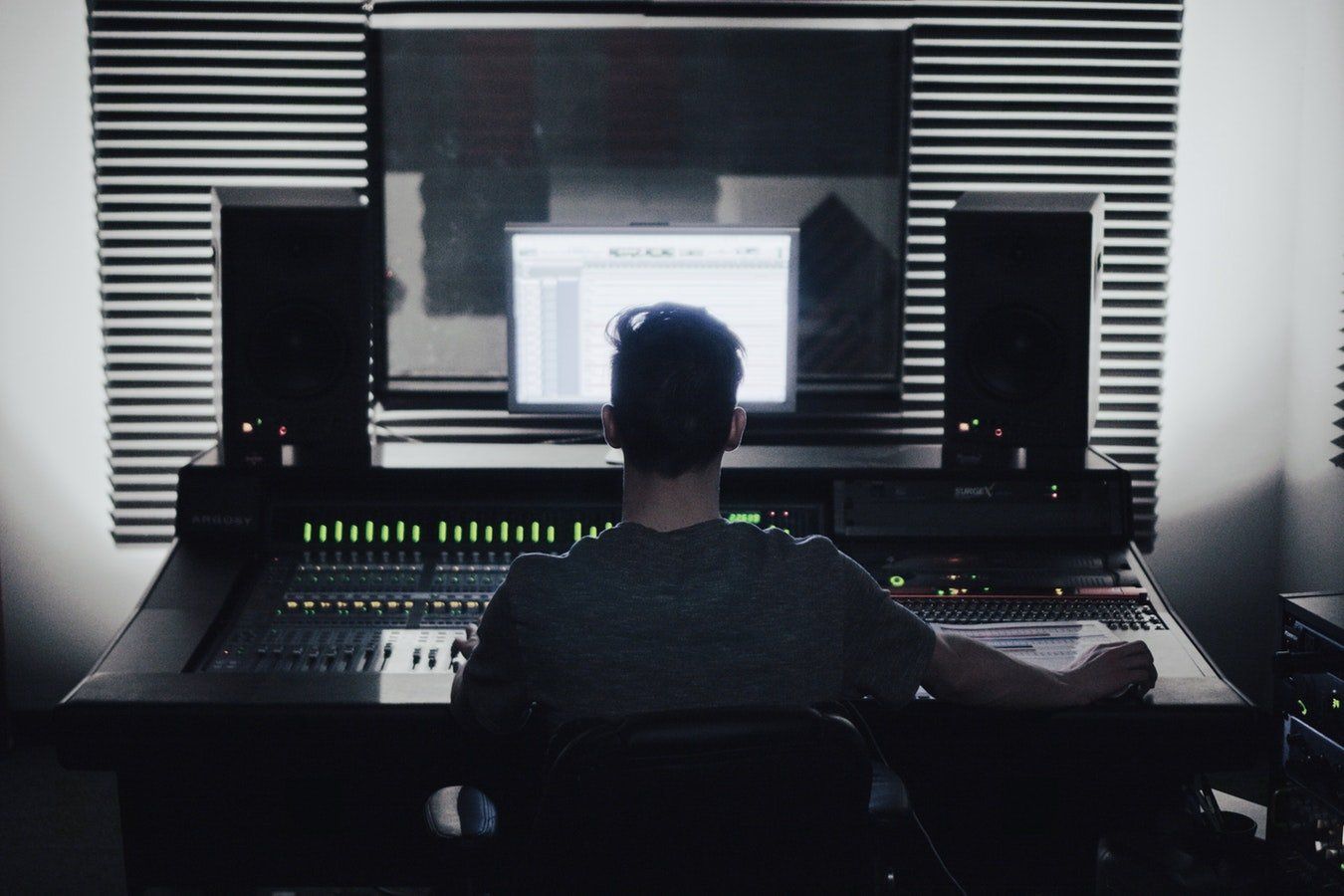 Person sits at audio mixing console in a studio, facing a computer screen.