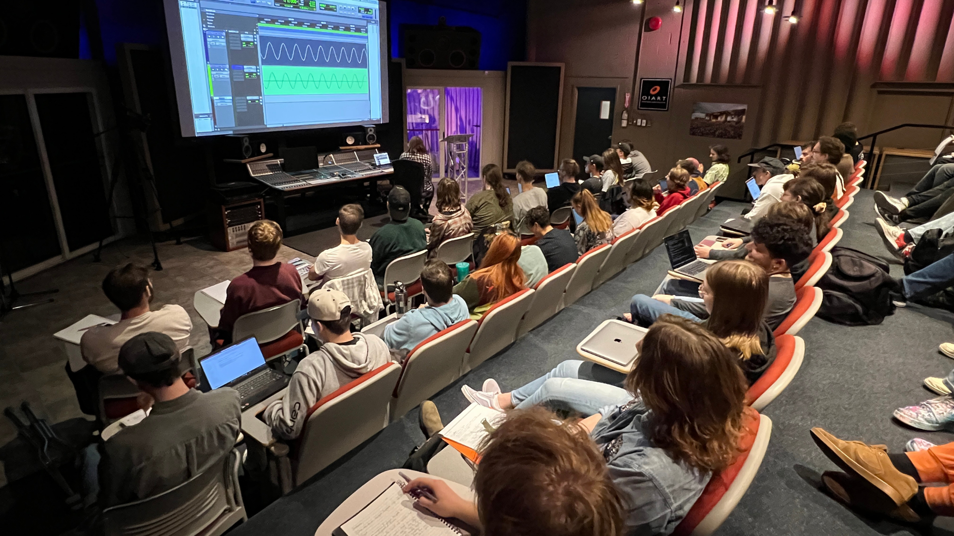 Students seated in a tiered classroom facing a large projector screen showing audio production software.