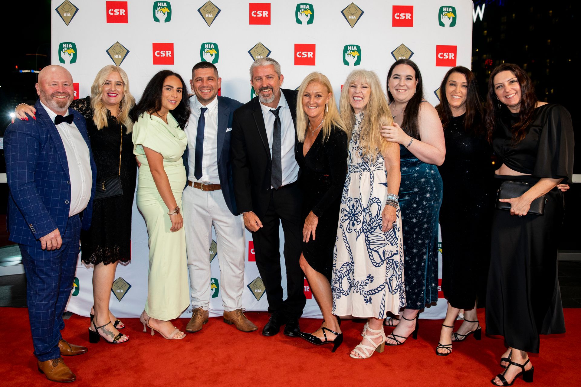 A group of ten people posing on a red carpet at an event. The group is smiling, some with arms around each other, against a branded backdrop — Evolution Building Group Dapto, NSW
