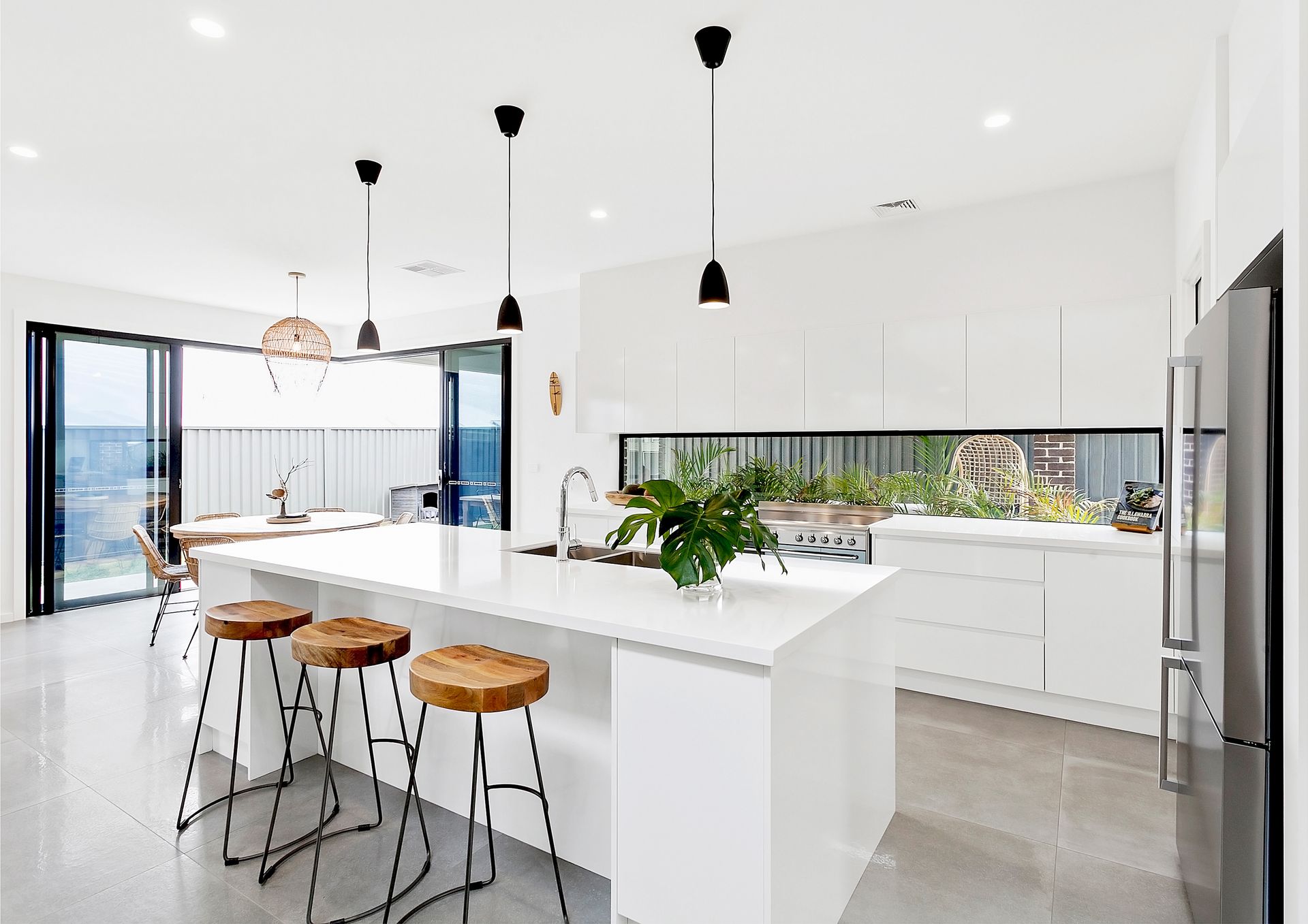 Modern white kitchen with island, stools, and black pendant lights. Sliding glass doors lead to an outdoor dining area — Evolution Building Group Dapto, NSW