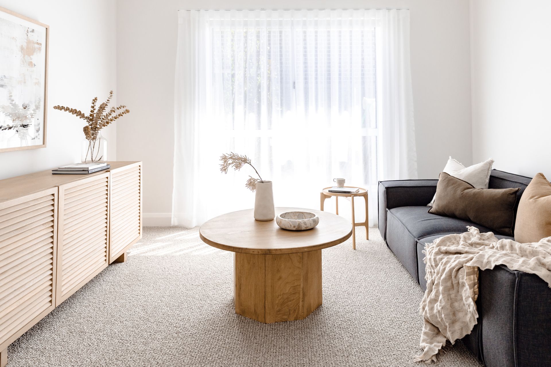 A bright living room with a grey sofa, wooden coffee table, and a light-coloured cabinet. Sheer curtains cover the large window — Evolution Building Group Dapto, NSW