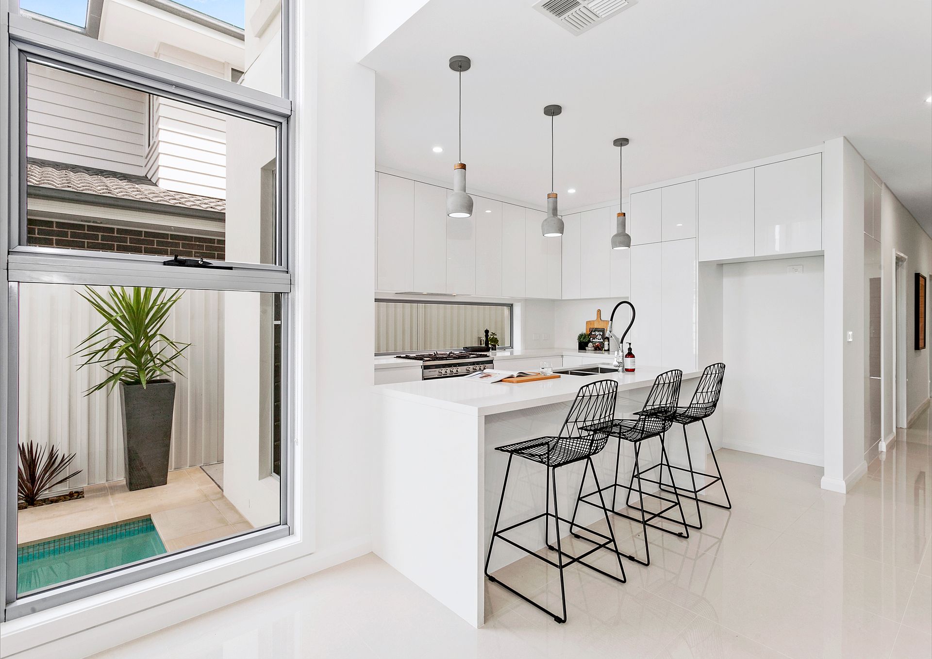 Bright white kitchen with a central island and three black wire-framed bar stools, a large window with a view to a small courtyard — Evolution Building Group Dapto, NSW