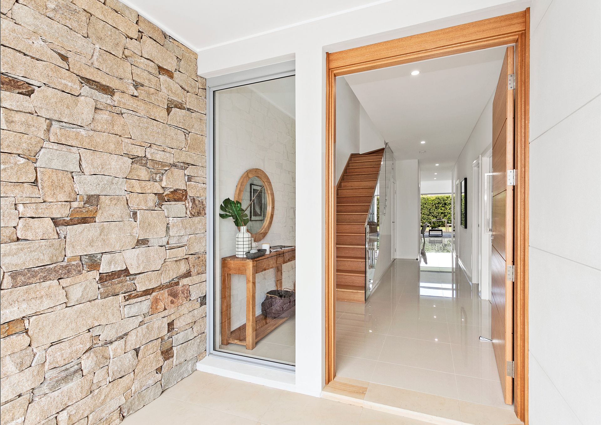 Entryway with stone wall, light wood door, and a hallway with a staircase and white walls. A mirrored table and plant are visible in the adjacent space — Evolution Building Group Dapto, NSW