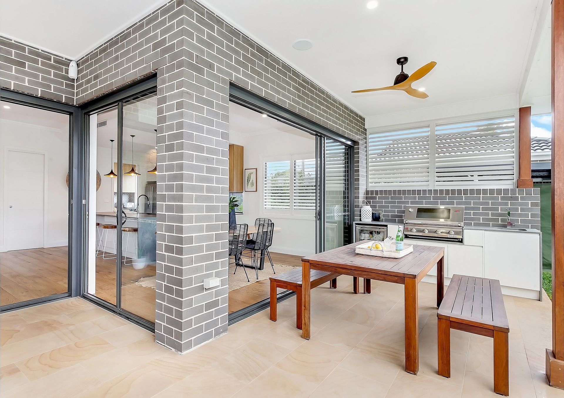 Outdoor patio with a wooden table and bench, looking into a modern kitchen. Grey brick walls and a ceiling fan are visible — Evolution Building Group In Shellharbour, NSW