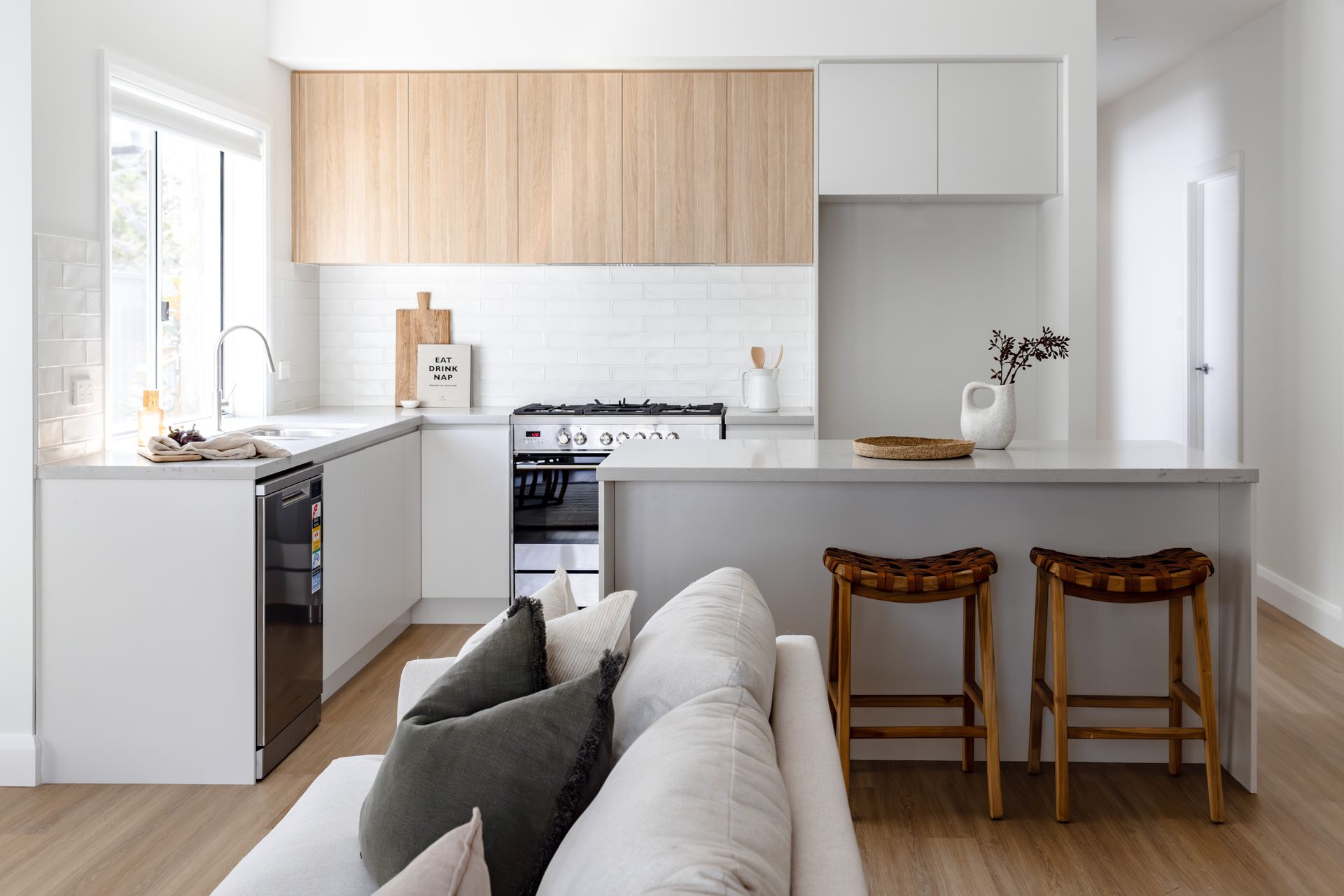 Modern kitchen with light wood cabinets, white countertops, and a grey island with two stools. A white couch is in front — Evolution Building Group In Shellharbour, NSW