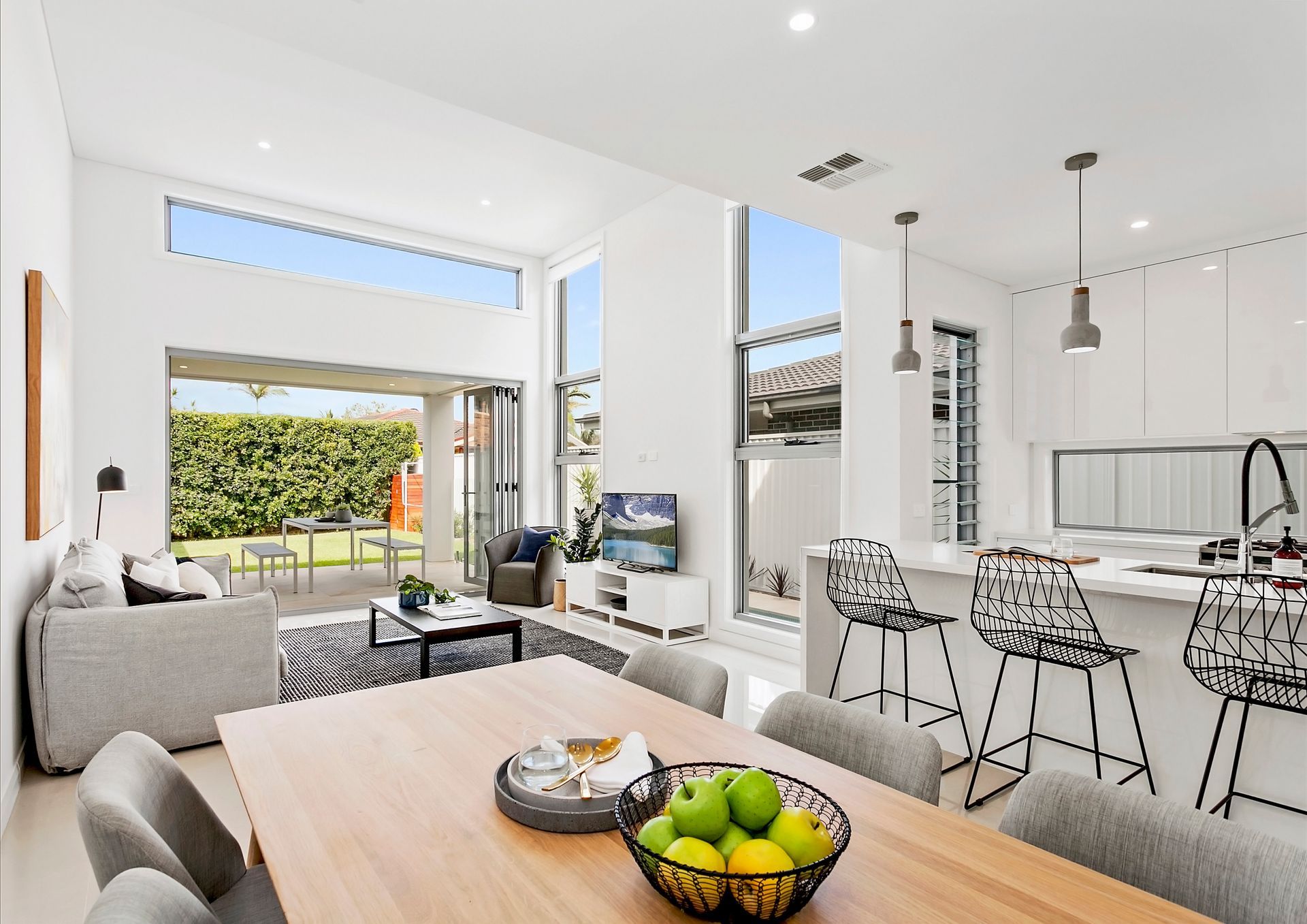 Modern open-plan living space with a kitchen, dining area, and living room that opens onto a backyard. Includes a dining table with fruit, a TV, and a sofa — Evolution Building Group In Berry, NSW