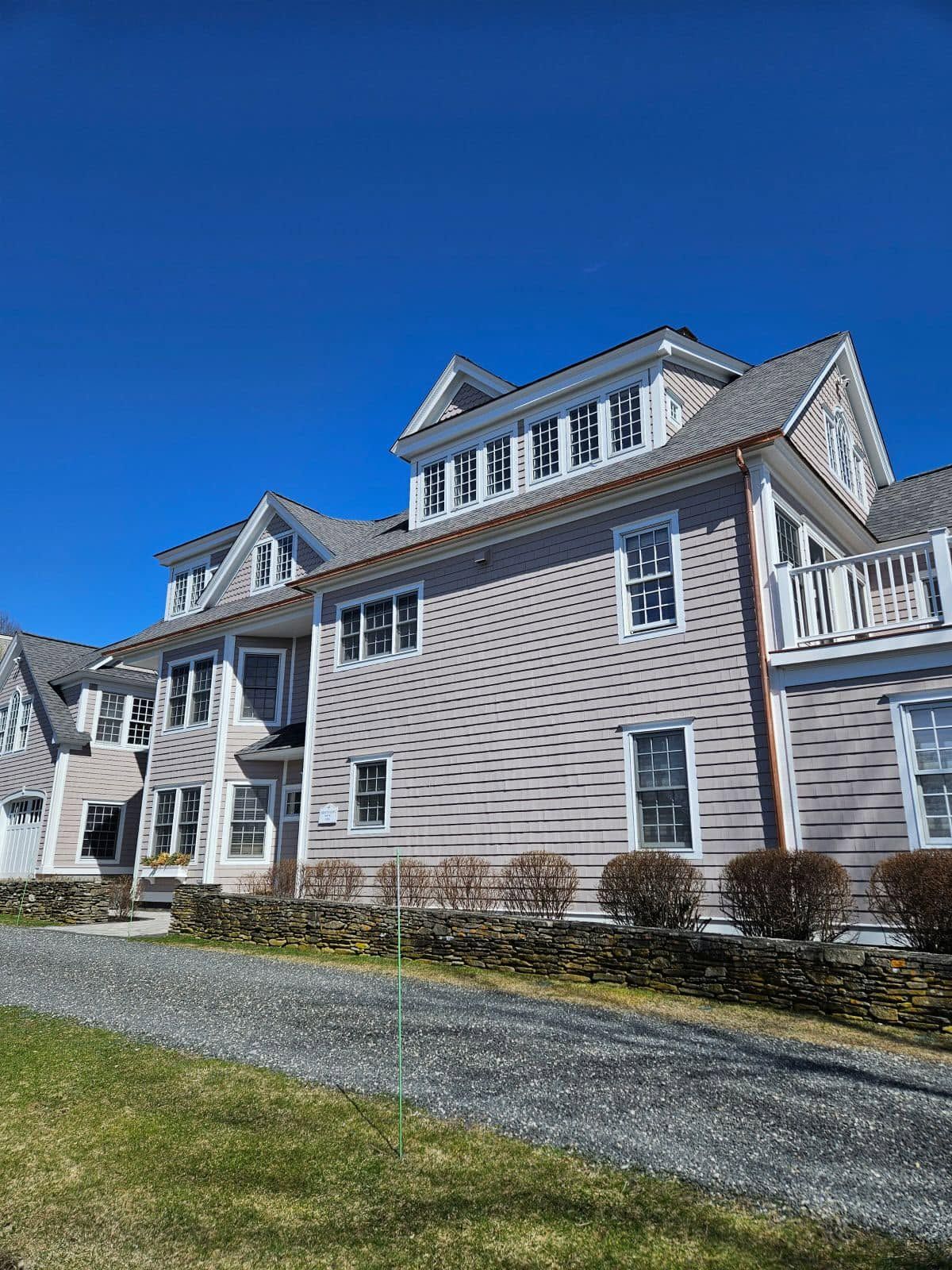 Large tan beach-style house with white trim under a clear blue sky, viewed from the lawn and driveway