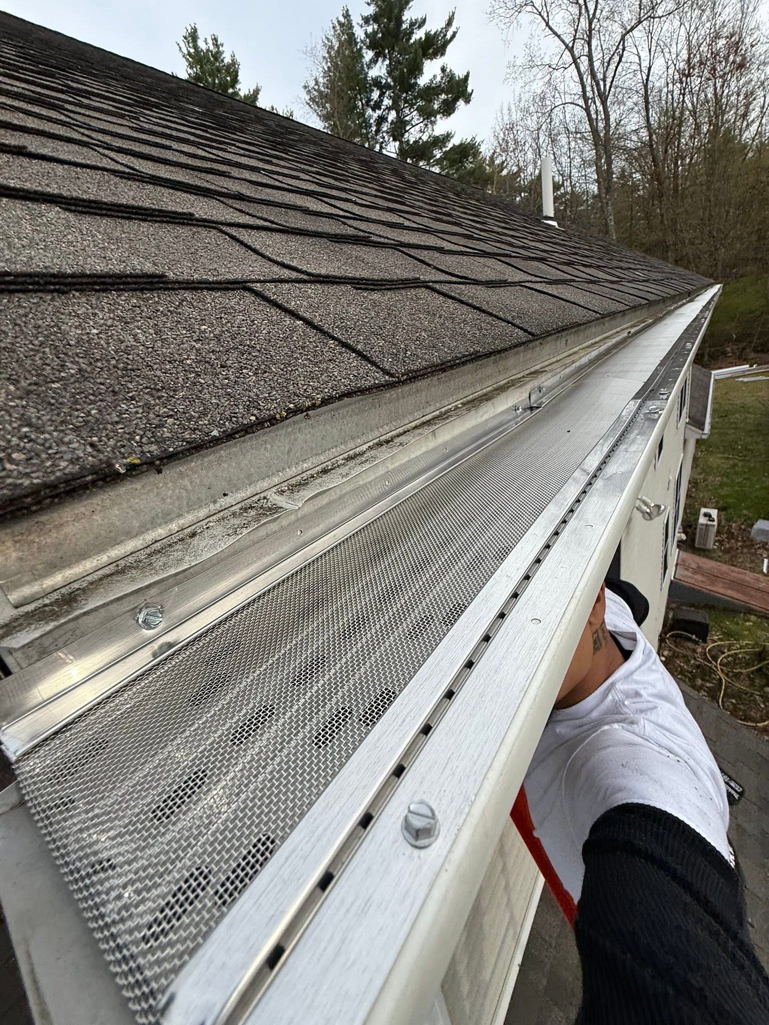 Person cleaning a white gutter along a shingled roof with trees in the background