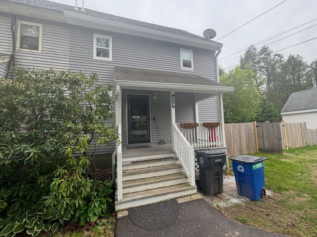 Two-story gray house with front steps, porch, and trash bins beside a fenced yard.