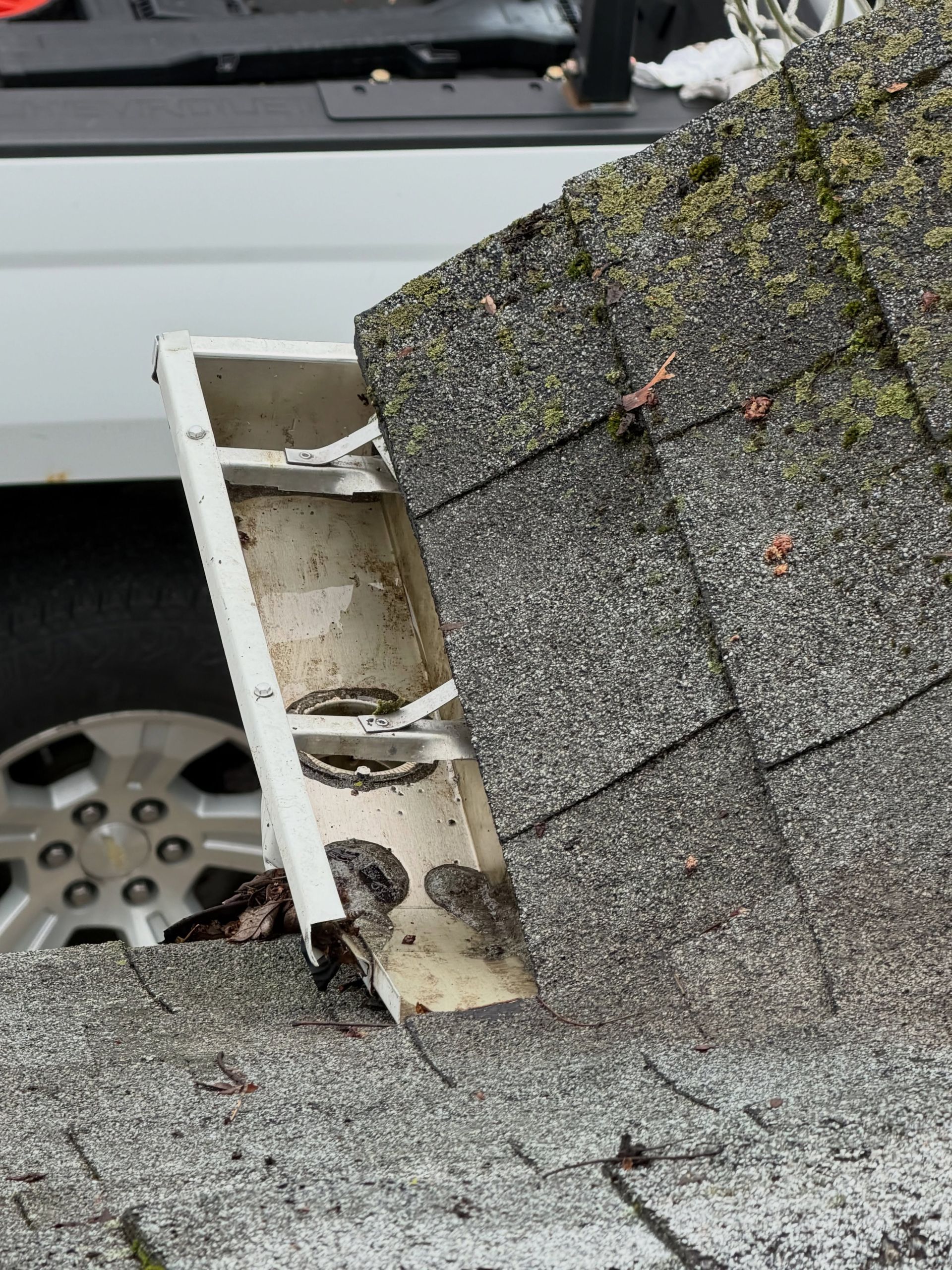 Damaged white gutter and roof shingles on a house, with debris and a vehicle wheel visible nearby
