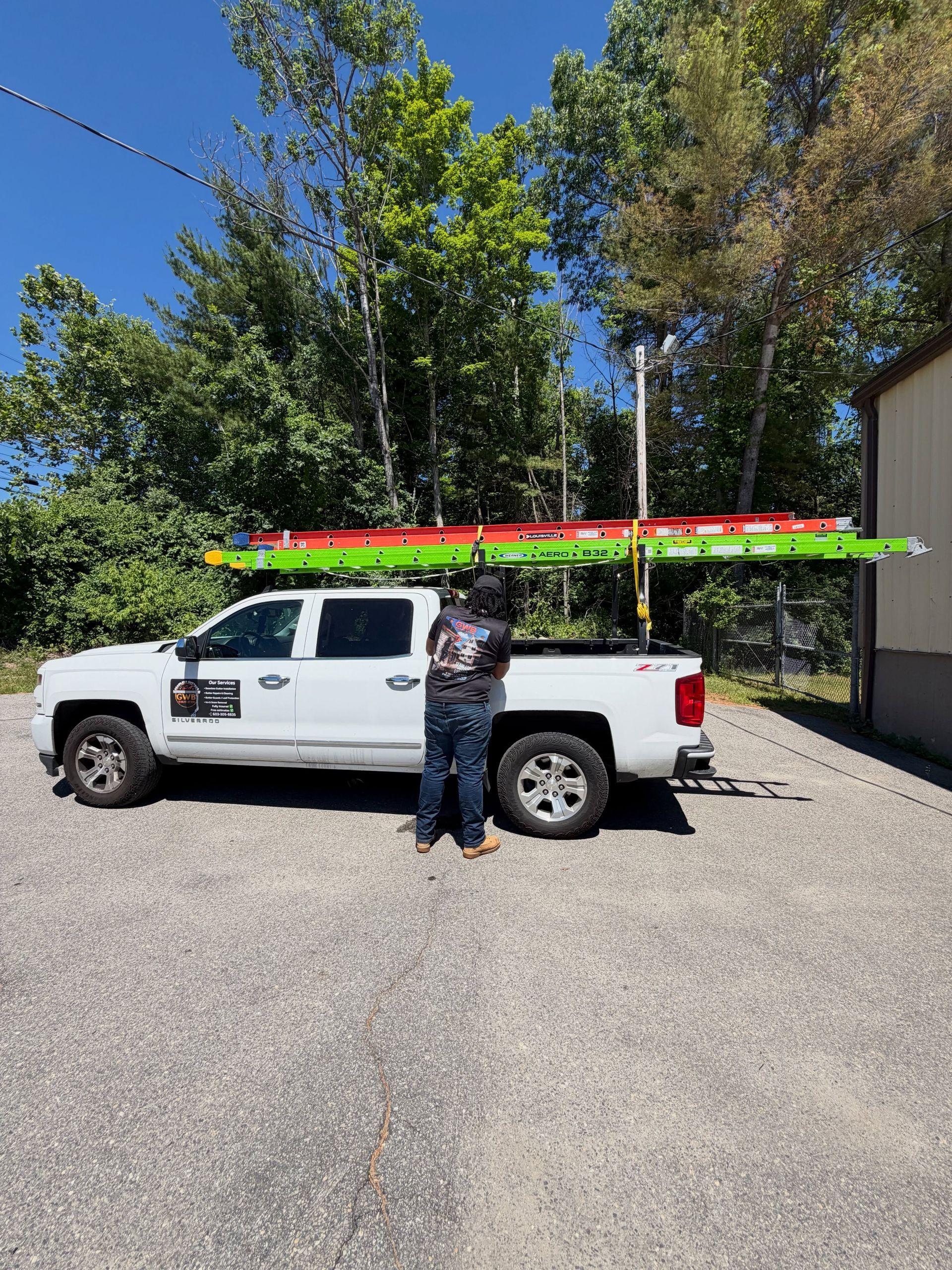 Person standing by a white pickup truck with colorful kayaks on top in a sunny gravel driveway