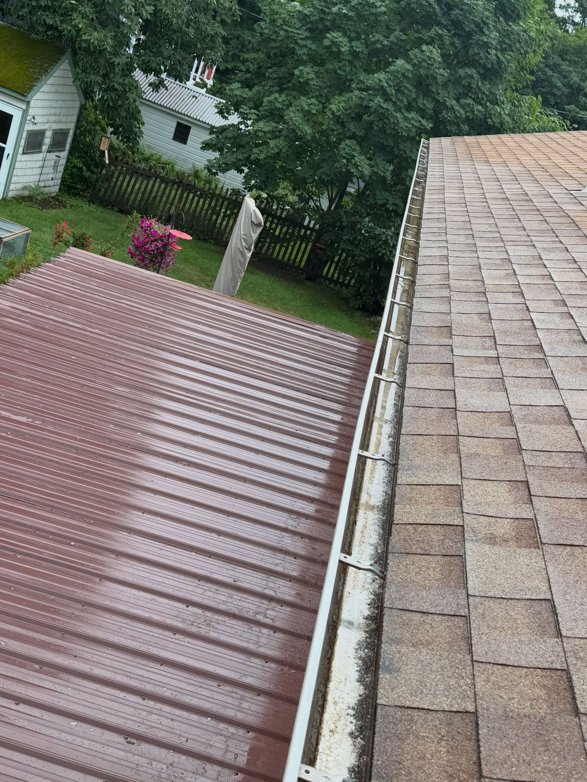 Roof shingles and a red metal awning beside a leafy backyard with neighboring houses.