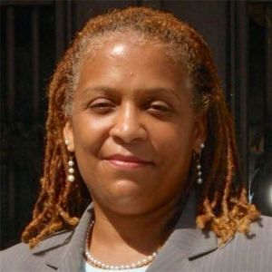 Woman with reddish-brown dreadlocks, smiling, wearing a blazer, pearl necklace, and earrings.