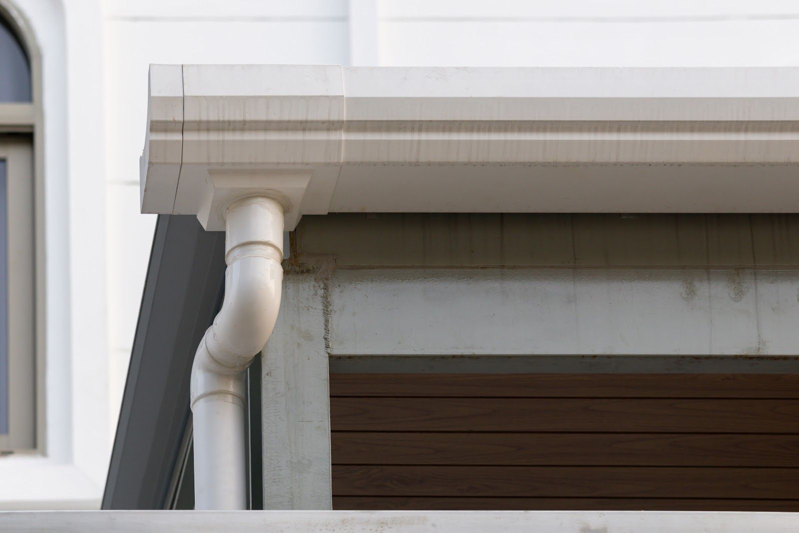 White house gutter and downspout beside a window and brown garage door under a roof eave