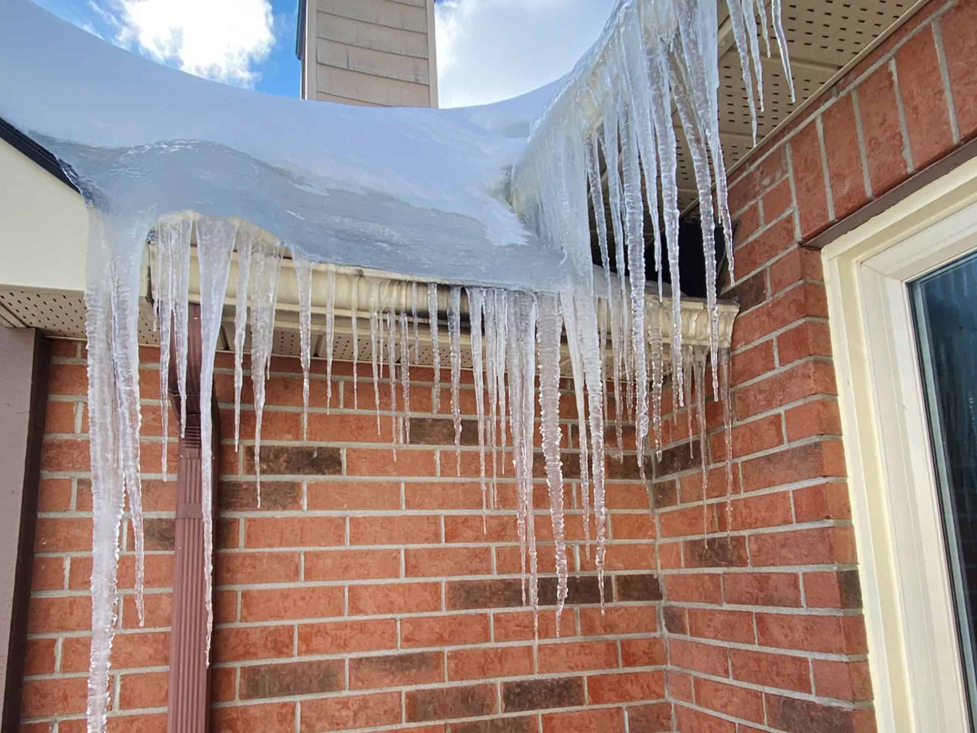 Icicles hang from a roof's edge above a brick wall and window on a sunny day.