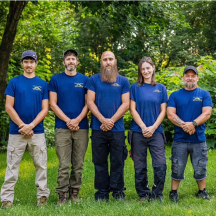 Five people in blue shirts stand on grass in front of trees. They appear to be a work crew.