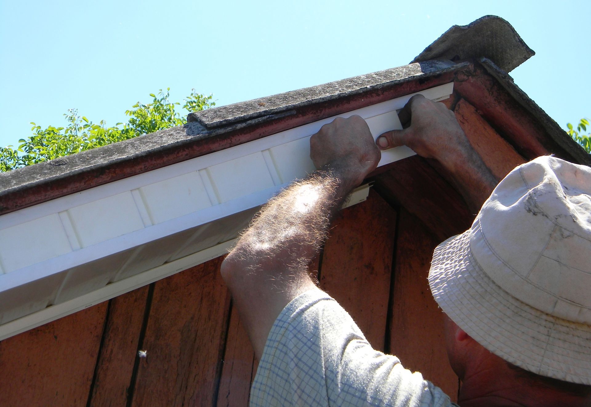 Person installing white siding on a wooden shed's edge, under a blue sky.