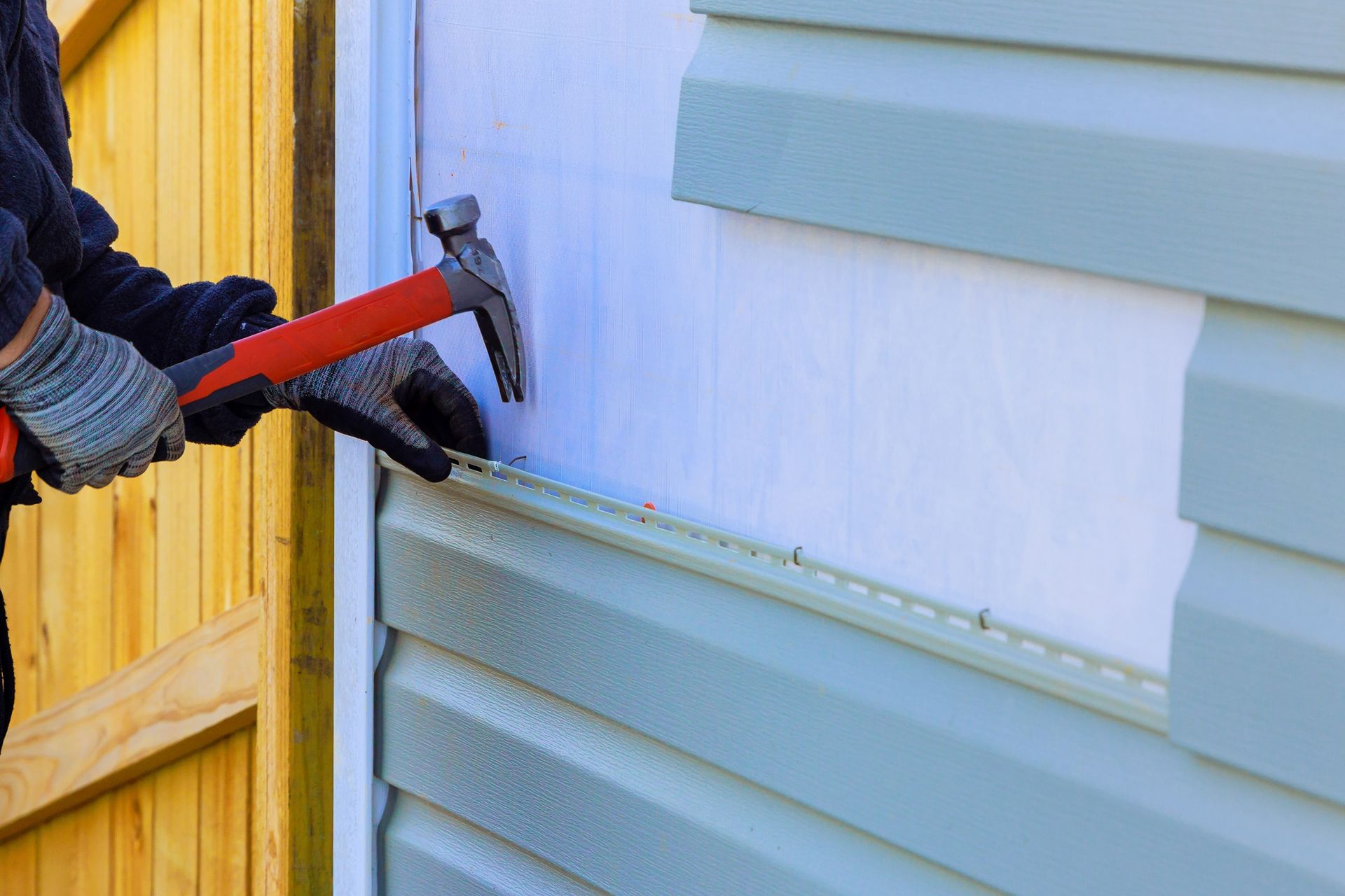 Person hammering light blue vinyl siding on a building, wearing gloves.