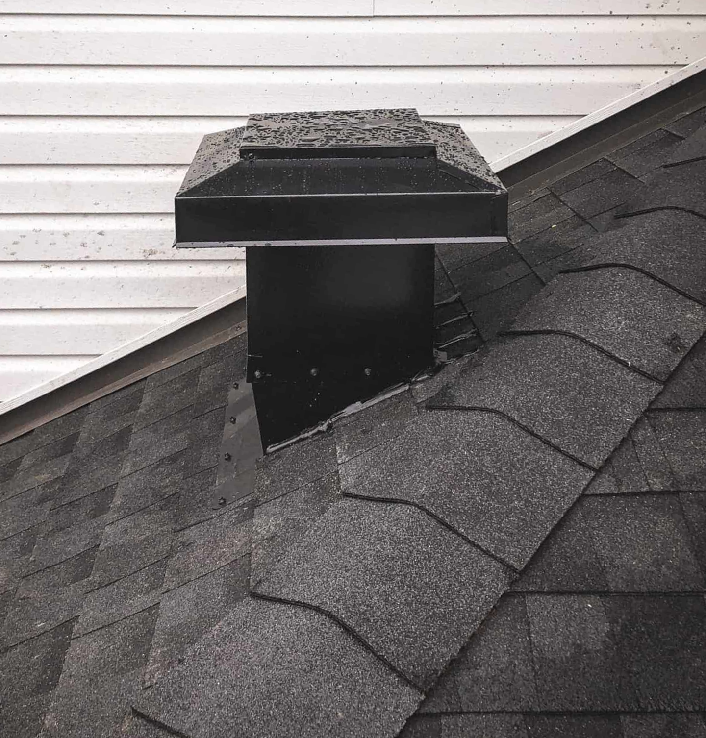 Black roof vent on a dark gray shingle roof, mounted near white siding.