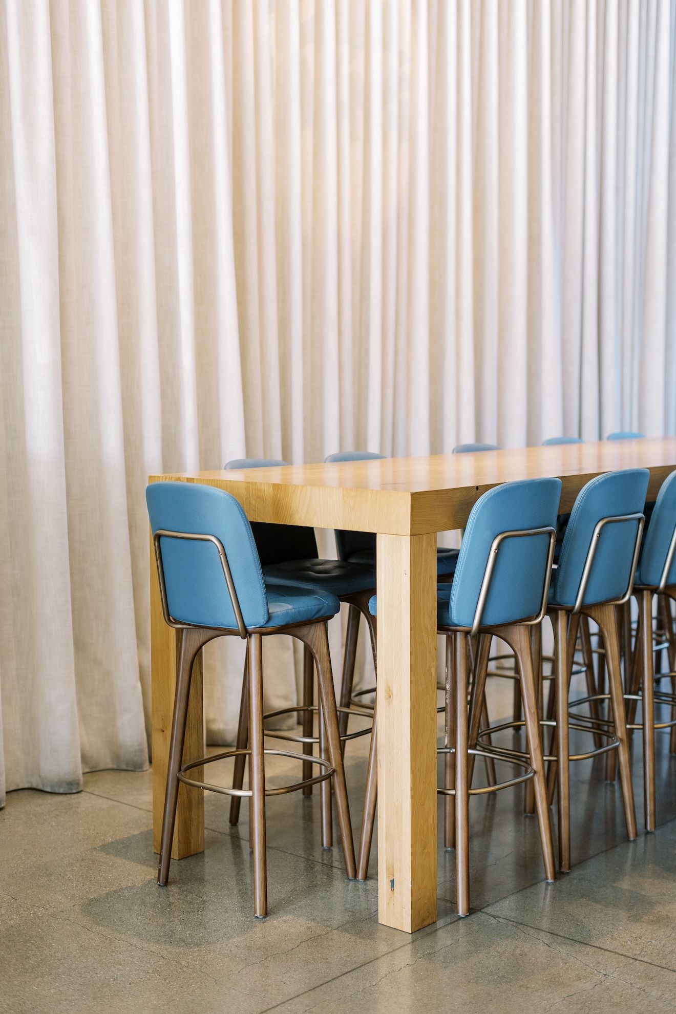 A long wooden table with blue chairs in front of a white curtain.