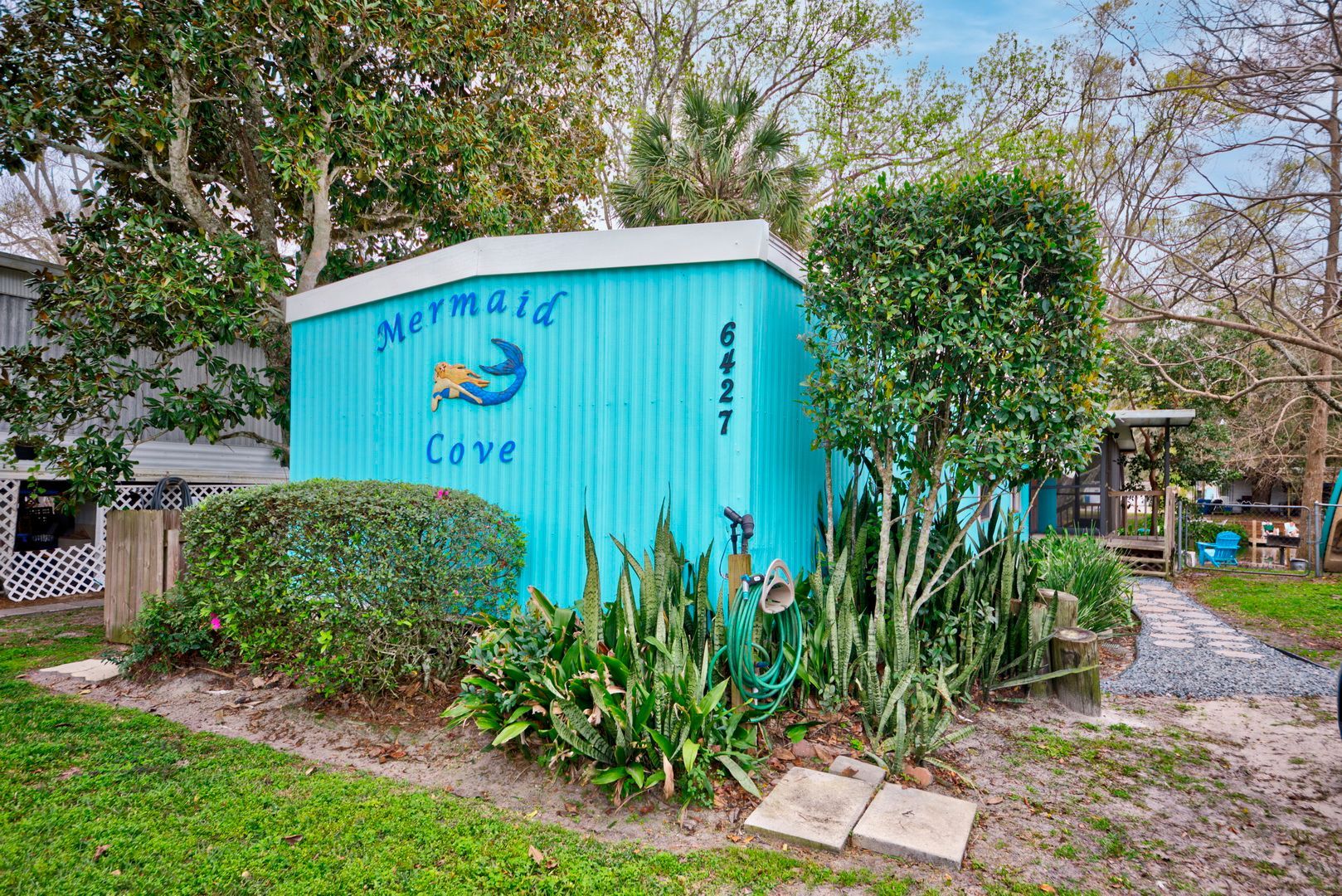 A blue shed with a mermaid on it is surrounded by trees and bushes.